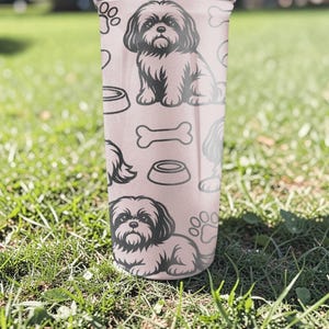 May include: A pink tumbler featuring a repeating pattern of a Shih Tzu dog, paw prints, dog bowls, and bones. The tumbler is cylindrical and appears to be made of a durable material. The background is blurred, showing green grass.