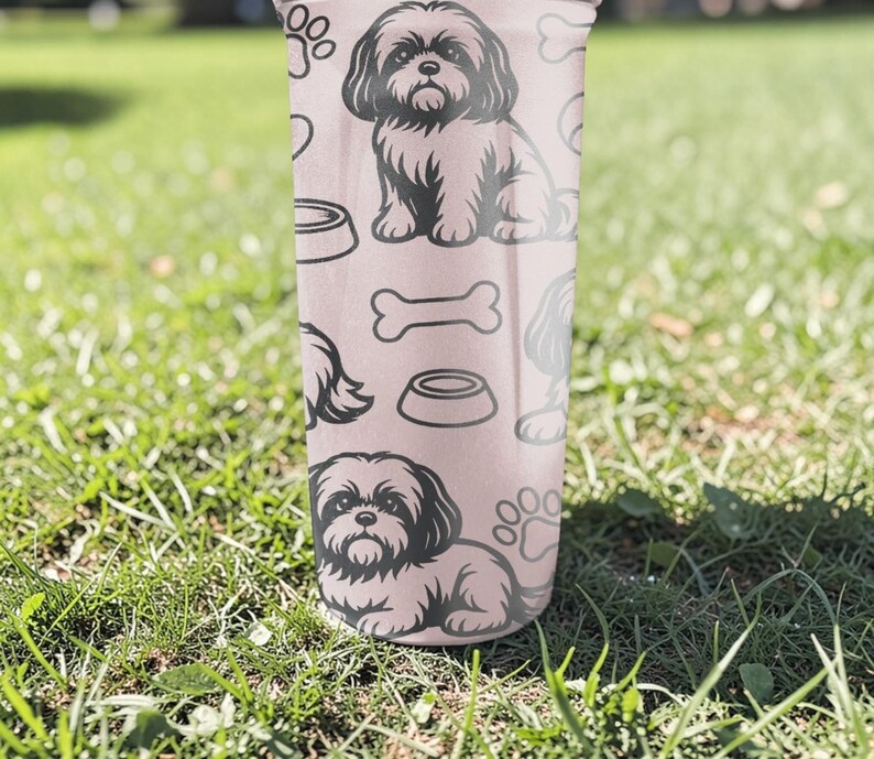 May include: A pink tumbler featuring a repeating pattern of a Shih Tzu dog, paw prints, dog bowls, and bones. The tumbler is cylindrical and appears to be made of a durable material. The background is blurred, showing green grass.
