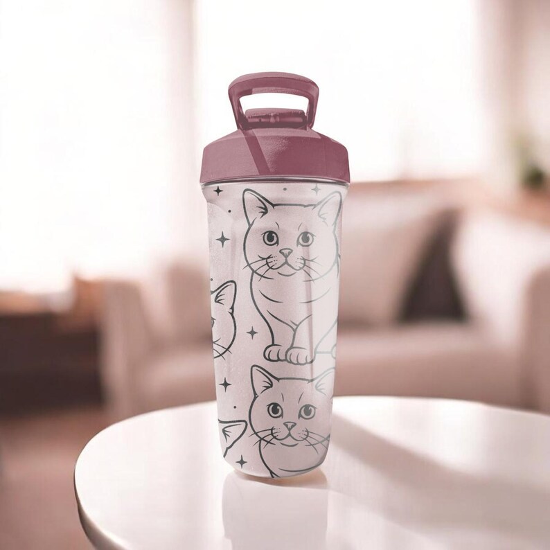 May include: A clear plastic water bottle with a maroon lid and handle. The bottle is decorated with a repeating pattern of cartoon cat faces and small star shapes. The bottle is sitting on a white table.