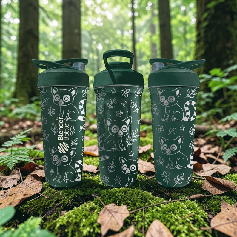 May include: Three green Blender Bottles with a lemur and floral design. The bottles have a dark green lid and a clear, flip-top opening. The lemur design is white and the bottles are sitting on a bed of moss and leaves.