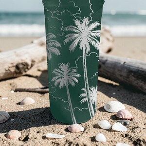 Może przedstawiać: Zielony kubek z białym wzorem palm i chmur. Cylindryczny kubek stoi na piaszczystej plaży, z muszlami i drewnem wyrzuconym na brzeg. W tle widoczne jest morze.