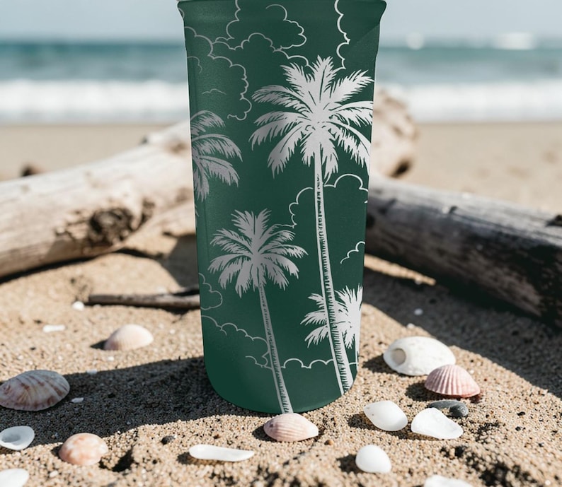 Może przedstawiać: Zielony kubek z białym wzorem palm i chmur. Cylindryczny kubek stoi na piaszczystej plaży, z muszlami i drewnem wyrzuconym na brzeg. W tle widoczne jest morze.