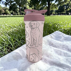 以下が含まれることがあります： フリップトップの蓋が付いた、薄いピンク色のウォーターボトル。ボトルには、グレーの犬のイラストが繰り返されるパターンがあります。ボトルは白い布の上に置かれ、草の背景があります。