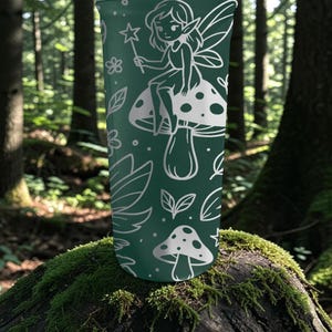 Puede incluir: Un vaso verde oscuro con una ilustraci&oacute;n blanca de un hada sentada en una seta, sosteniendo una varita m&aacute;gica. El dise&ntilde;o incluye hojas y flores. El vaso es cil&iacute;ndrico y parece ser de metal.