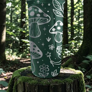 Puede incluir: Un vaso verde oscuro con un dise&ntilde;o caprichoso de setas blancas, estrellas, flores y un hada con una varita m&aacute;gica. El vaso cil&iacute;ndrico est&aacute; sobre un toc&oacute;n cubierto de musgo, con un fondo de bosque.