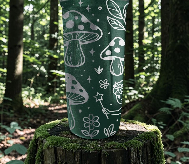 Puede incluir: Un vaso verde oscuro con un dise&ntilde;o caprichoso de setas blancas, estrellas, flores y un hada con una varita m&aacute;gica. El vaso cil&iacute;ndrico est&aacute; sobre un toc&oacute;n cubierto de musgo, con un fondo de bosque.