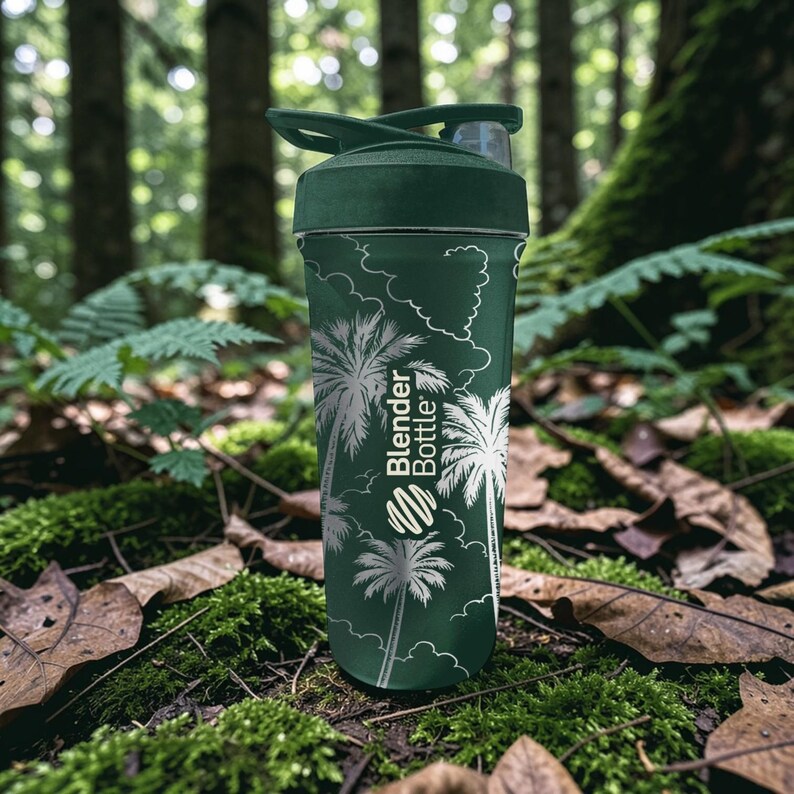 Może przedstawiać: Zielona butelka Blender Bottle z motywem palm. Butelka ma ciemnozieloną pokrywę i biały wz&oacute;r palm i chmur na korpusie. Butelka stoi na zielonym mchu i opadłych liściach.