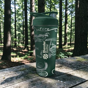 May include: A dark green Blender Bottle with a golf-themed design. The bottle features white illustrations of golf clubs, a golf cart, and a golf ball. The words "fore", "Mulligan", and "Bunker" are also visible on the bottle.