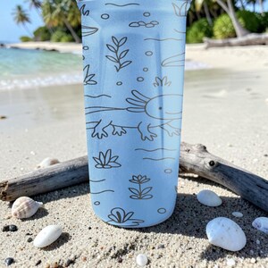 May include: A light blue tumbler with a brown axolotl and aquatic plant design. The tumbler is cylindrical and features a detailed illustration of an axolotl and aquatic plants. The design is printed on the tumbler's surface.