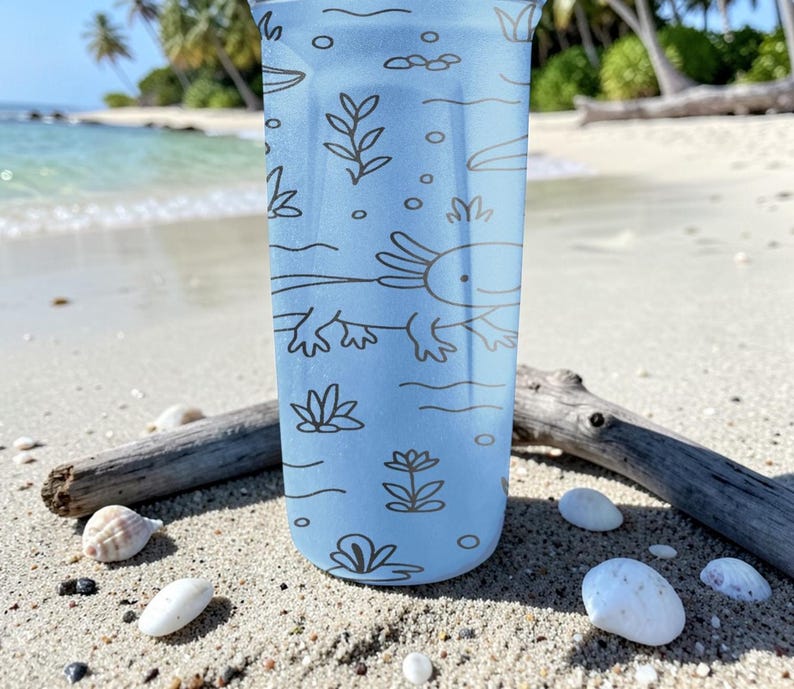 May include: A light blue tumbler with a brown axolotl and aquatic plant design. The tumbler is cylindrical and features a detailed illustration of an axolotl and aquatic plants. The design is printed on the tumbler's surface.