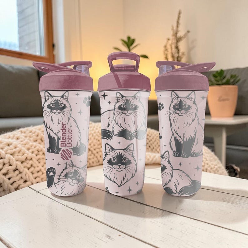 May include: Three Blender Bottles with a light pink base and a dark pink lid. Each bottle features a black and white illustration of a fluffy cat, paw prints, and stars. The text "Blender Bottle" is printed on the side of one bottle.