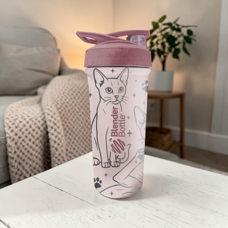 Puede incluir: Una botella Blender rosa con un dise&ntilde;o de gato. La botella tiene una tapa rosa y un cuerpo transparente con una ilustraci&oacute;n de gato, huellas de patas y estrellas. Las palabras "Blender Bottle" est&aacute;n impresas en el lateral.
