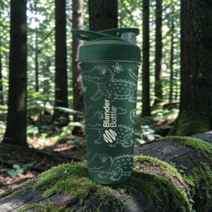 Peut inclure: Une gourde verte Blender Bottle avec un couvercle rabattable, orn&eacute;e d'un motif d'alligator blanc. La bouteille est cylindrique et repose sur une b&ucirc;che couverte de mousse dans une for&ecirc;t. Le logo Blender Bottle est visible.