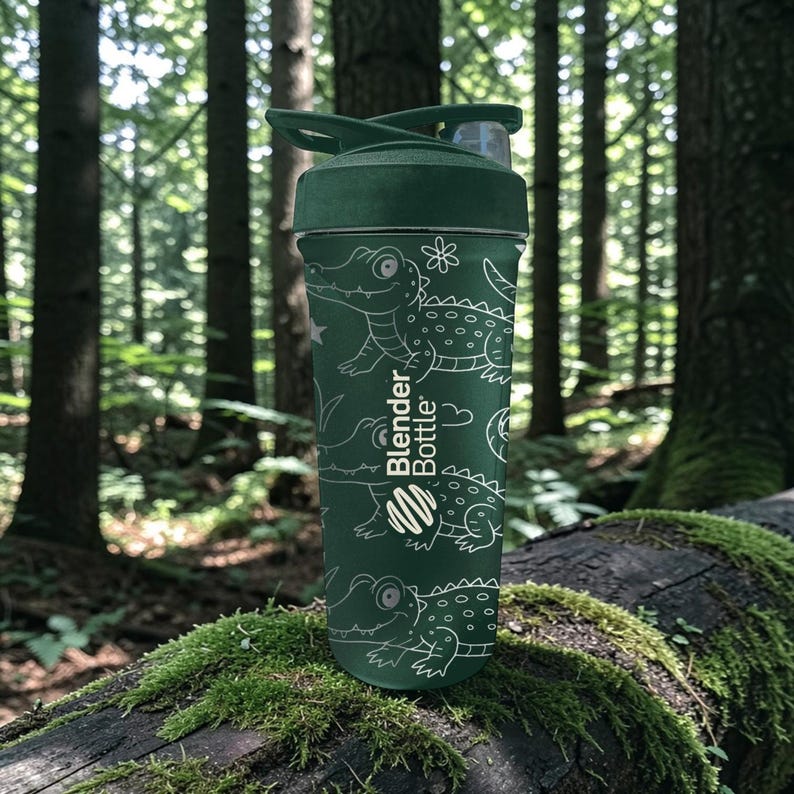 Peut inclure: Une gourde verte Blender Bottle avec un couvercle rabattable, orn&eacute;e d'un motif d'alligator blanc. La bouteille est cylindrique et repose sur une b&ucirc;che couverte de mousse dans une for&ecirc;t. Le logo Blender Bottle est visible.