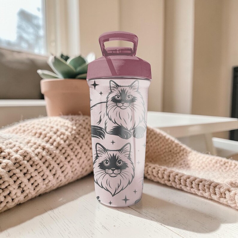 May include: A light pink water bottle with a dark pink lid and handle. The bottle features a black illustration of a fluffy cat and stars. The cat illustration is repeated twice on the bottle.