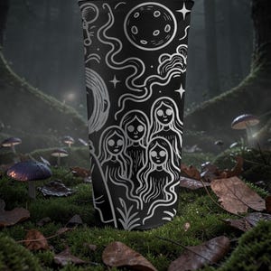 May include: A black tumbler with a white illustration of celestial elements and figures. The design includes a moon, stars, and stylized women. The tumbler is cylindrical and stands upright against a forest backdrop with mushrooms and foliage.