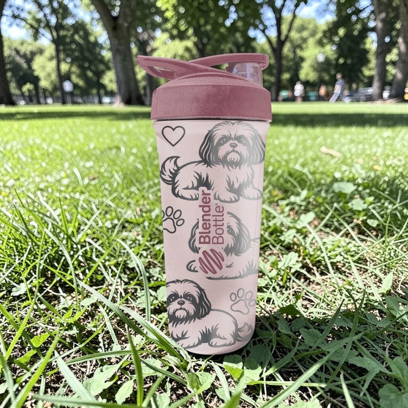May include: A pink Blender Bottle with a matching lid, featuring a heart, paw prints, and illustrations of a Shih Tzu dog. The bottle is cylindrical and sits on a grassy surface.