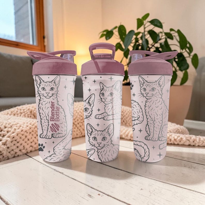 May include: Three white Blender Bottles with a pink lid and handle. The bottles feature a repeating pattern of cat illustrations in black and white, with stars and paw prints. The text "Blender Bottle" is printed on one bottle.
