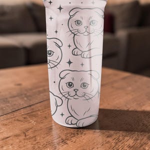 May include: A light pink tumbler featuring a repeating pattern of cartoon Scottish Fold cats in black outline. The cats are in a sitting position with their ears folded. Small black star shapes are scattered around the cats.