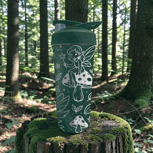 Puede incluir: Una botella de agua verde oscuro con un dise&ntilde;o de hada y hongo blanco. La botella tiene una tapa abatible y es cil&iacute;ndrica. El hada est&aacute; sentada en un hongo y sosteniendo una varita en forma de estrella. El fondo es un bosque.