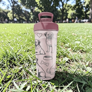 May include: A pink water bottle with a matching lid and handle. The bottle features line drawings of a pug, a dog bowl, a bone, and a heart. The bottle is sitting on green grass.