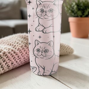 May include: A pink tumbler with two cartoon cat illustrations and star accents. The cats are drawn in black outline, with detailed facial features. The tumbler is cylindrical and appears to be made of a durable material.