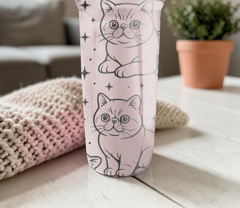 May include: A pink tumbler with two cartoon cat illustrations and star accents. The cats are drawn in black outline, with detailed facial features. The tumbler is cylindrical and appears to be made of a durable material.