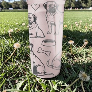 May include: A light pink tumbler featuring a repeating pattern of dog illustrations, a heart, a dog bowl, and a bone. The tumbler is cylindrical and appears to be made of a durable material. The dog illustrations are in a simple, line-art style.