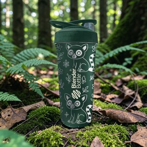 May include: A dark green Blender Bottle with a white lemur and floral pattern. The bottle has a dark green lid and a flip-top closure. The words "Blender Bottle" are printed on the side. The bottle is set against a forest background.