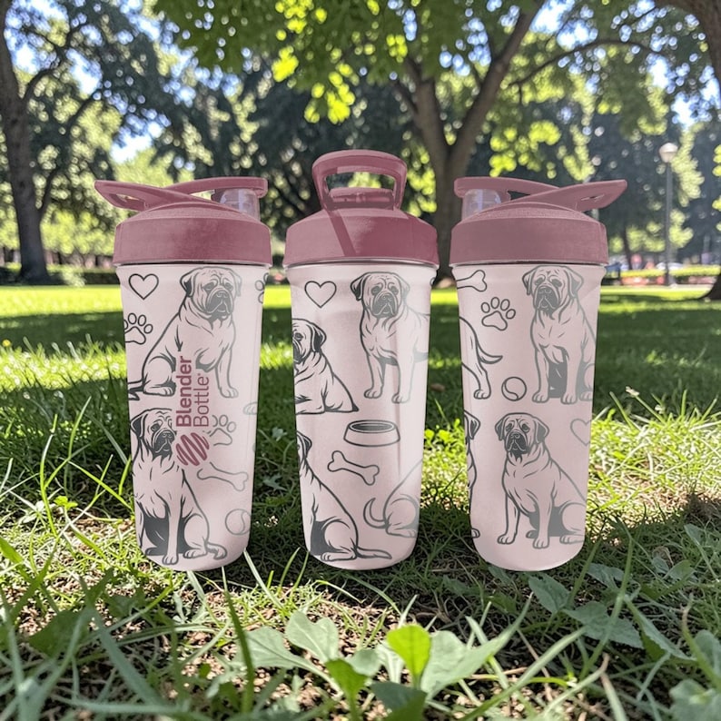 May include: Three pink Blender Bottles with maroon lids and handles, each adorned with a repeating pattern of dog illustrations, hearts, paw prints, and bones. The bottles are set on green grass.