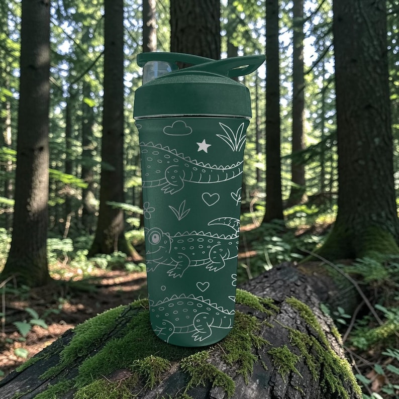 Peut inclure: Une gourde verte avec un couvercle rabattable, orn&eacute;e de dessins au trait blancs d'alligators, de nuages, d'&eacute;toiles et de c&oelig;urs. La gourde est plac&eacute;e devant un fond de for&ecirc;t avec de grands arbres et des rondins couverts de mousse.