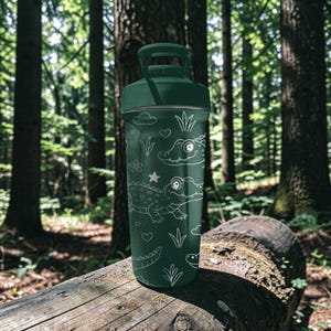 Peut inclure: Une gourde vert fonc&eacute; avec un motif d'alligator blanc. La gourde a une poign&eacute;e et un couvercle vissable. Le motif comprend des alligators de dessins anim&eacute;s, des &eacute;toiles, des c&oelig;urs et des plantes. La gourde est pos&eacute;e sur une b&ucirc;che dans une for&ecirc;t.