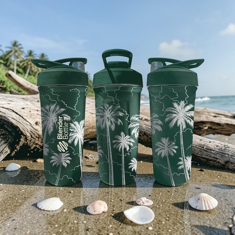 Może przedstawiać: Trzy zielone butelki Blender Bottle z białymi wzorami palm stoją na piaszczystej plaży. Butelki mają ciemnozielone pokrywki i uchwyty. Logo Blender Bottle jest widoczne na jednej butelce. Muszle są rozrzucone na piasku.