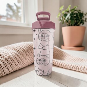 May include: A pink and clear water bottle with two cartoon cat illustrations and star accents. The bottle has a dark pink lid and handle. The cat illustrations are in a line art style.