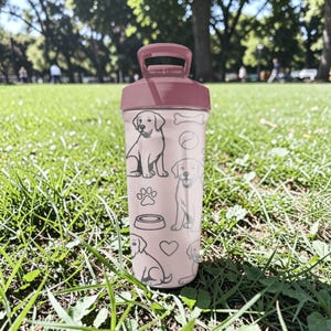May include: A pink water bottle with a maroon lid and handle. The bottle is decorated with line drawings of dogs, paw prints, a dog bowl, a bone, and a heart. The bottle is sitting on a grassy surface.