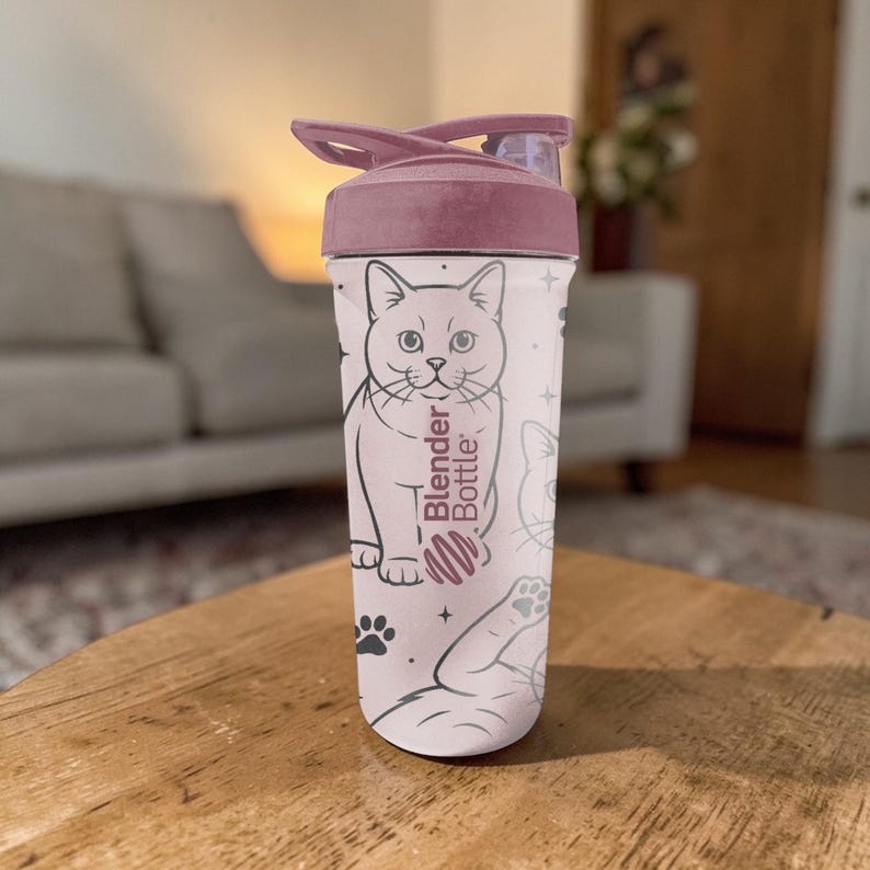 May include: A Blender Bottle with a pink lid and a light pink body. The body features a repeating pattern of cat illustrations, paw prints, and stars. The Blender Bottle logo is printed vertically on the side.