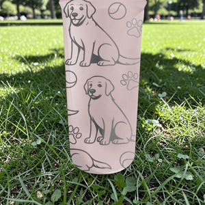 May include: A light pink tumbler with a repeating pattern of a dog, paw prints, and a ball. The dog is light grey and sitting. The cylindrical tumbler appears to be made of plastic.