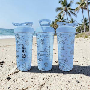 May include: Three light blue Blender Bottles with axolotl illustrations and the Blender Bottle logo. The bottles are on a sandy beach with the ocean and palm trees in the background. Each bottle has a flip-top lid.
