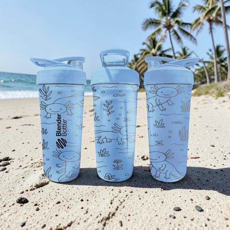 May include: Three light blue Blender Bottles with axolotl illustrations and the Blender Bottle logo. The bottles are on a sandy beach with the ocean and palm trees in the background. Each bottle has a flip-top lid.