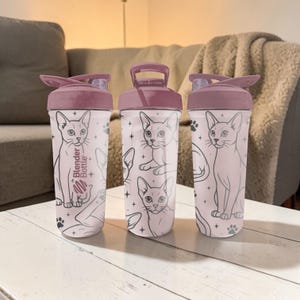 Puede incluir: Tres botellas Blender con tapa rosa y cuerpo rosa claro. Las botellas presentan un patr&oacute;n repetido de ilustraciones de gatos y huellas de patas. El texto "Blender Bottle" est&aacute; impreso en una botella.