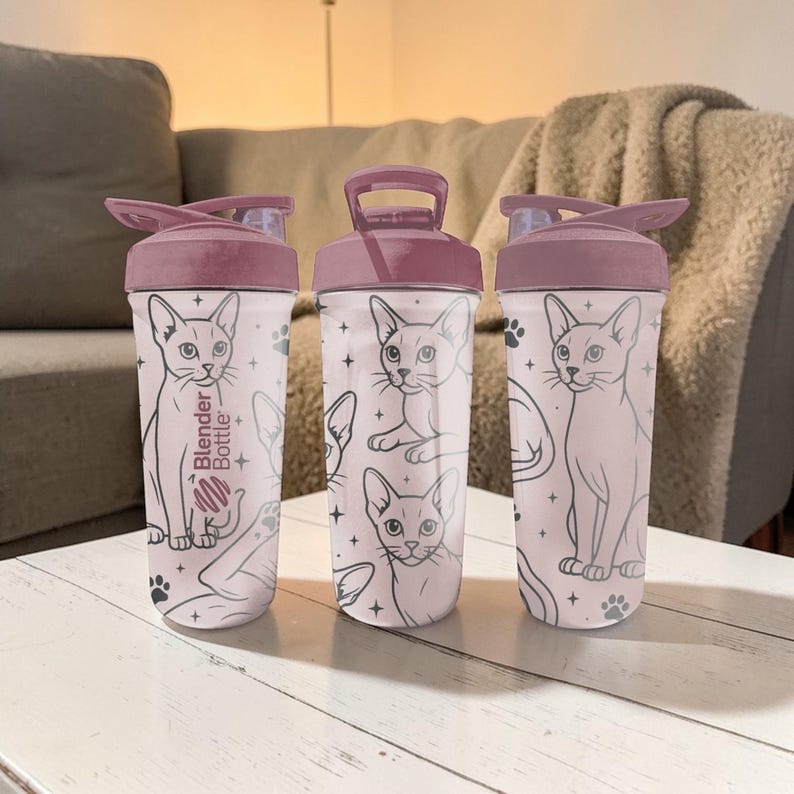Puede incluir: Tres botellas Blender con tapa rosa y cuerpo rosa claro. Las botellas presentan un patr&oacute;n repetido de ilustraciones de gatos y huellas de patas. El texto "Blender Bottle" est&aacute; impreso en una botella.
