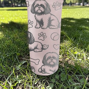 May include: A light pink tumbler featuring a Shih Tzu dog design. The tumbler has multiple illustrations of the dog, paw prints, a bone, a heart, and a ball. The tumbler is sitting on green grass.