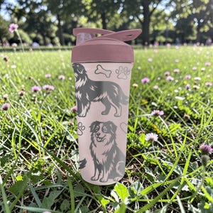 K&ouml;nnte beinhalten: Eine rosa Wasserflasche mit Klappdeckel, verziert mit grauen Illustrationen von Hunden, Pfotenabdr&uuml;cken und einem Knochen. Die Flasche befindet sich in einer grasbewachsenen Umgebung mit Blumen, ideal f&uuml;r Outdoor-Aktivit&auml;ten.