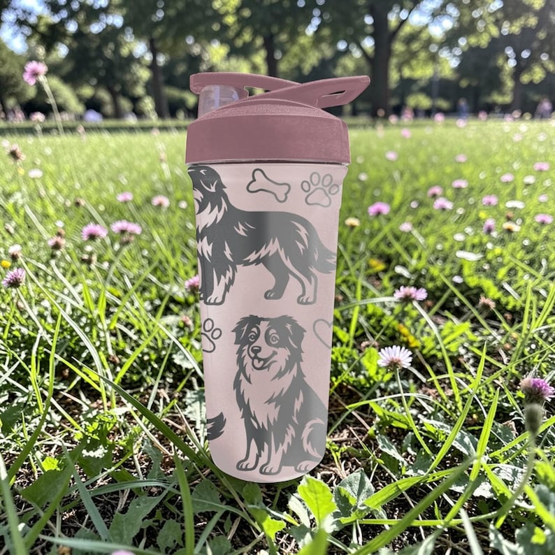 K&ouml;nnte beinhalten: Eine rosa Wasserflasche mit Klappdeckel, verziert mit grauen Illustrationen von Hunden, Pfotenabdr&uuml;cken und einem Knochen. Die Flasche befindet sich in einer grasbewachsenen Umgebung mit Blumen, ideal f&uuml;r Outdoor-Aktivit&auml;ten.