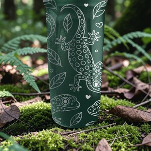May include: A dark green tumbler with a white gecko and leaf pattern. The design includes a large gecko, smaller gecko faces, leaves, and hearts. The tumbler is cylindrical and appears to be made of metal.