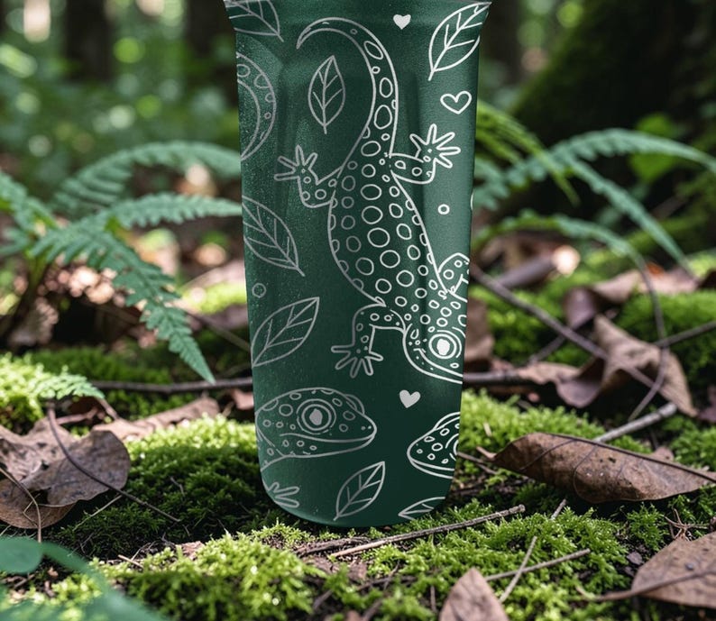 May include: A dark green tumbler with a white gecko and leaf pattern. The design includes a large gecko, smaller gecko faces, leaves, and hearts. The tumbler is cylindrical and appears to be made of metal.