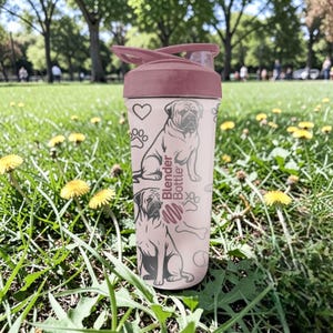 May include: A pink Blender Bottle with a maroon lid and a dog graphic. The bottle features a dog illustration, paw prints, and the words "Blender Bottle". The bottle is designed for mixing protein shakes or other beverages.