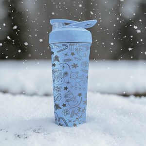 Puede incluir: Una botella de agua azul claro con tapa abatible, decorada con un dise&ntilde;o de vacaciones de invierno. El dise&ntilde;o incluye copos de nieve, mu&ntilde;ecos de nieve, renos y bastones de caramelo. La botella est&aacute; en un entorno nevado.