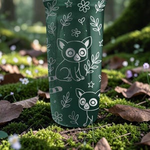 May include: A dark green tumbler with a white illustration of a cartoon lemur, leaves, flowers, and stars. The tumbler is cylindrical and appears to be made of metal or a similar material. The design is repeated around the tumbler.