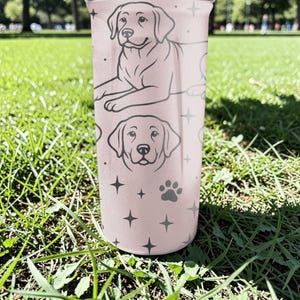 以下が含まれることがあります： ラブラドールレトリバーのイラストが描かれた、薄いピンク色のタンブラー。犬の足跡、星、2つの犬の肖像画が含まれています。円筒形のタンブラーは金属製のように見えます。